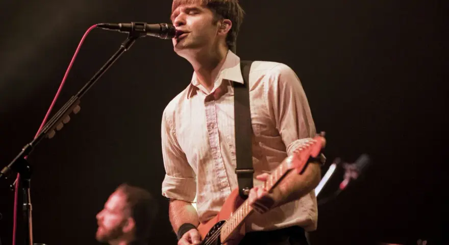 Ben Gibbard chante et joue de la guitare sur scène, éclairé par les projecteurs.