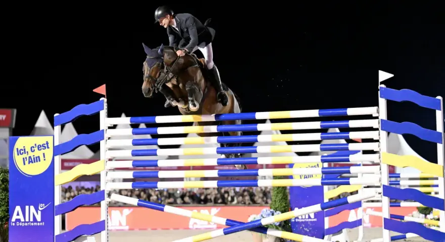 Cavalier et cheval en plein saut lors de l'épreuve des 6 barres. Le cheval est au sommet de son effort, les pattes repliées, franchissant l'obstacle.
