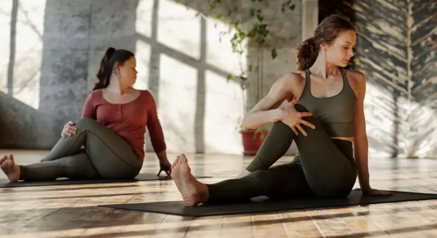 Dwie kobiety wykonują skręty tułowia na macie. Delikatne stretching poprawia samopoczucie.