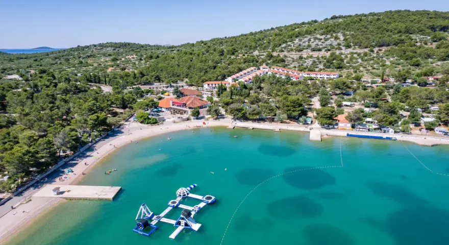 Malownicza zatoka z turkusową wodą, plażą i wodnym placem zabaw. W tle widać zabudowania i zielone wzgórza, idealne na camping Murter.