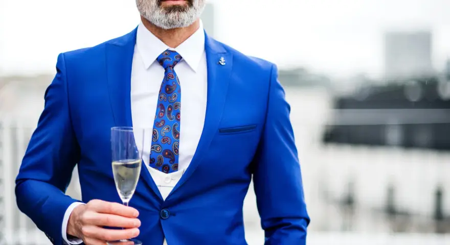 Elegancki mężczyzna w niebieskim garniturze i wzorzystym krawacie, trzymający kieliszek szampana. Idealny strój koktajlowy na specjalne okazje.
