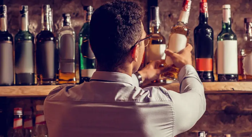 Mann wählt eine Flasche aus, in der reift Whisky in der Flasche nach. Viele Flaschen stehen im Regal.