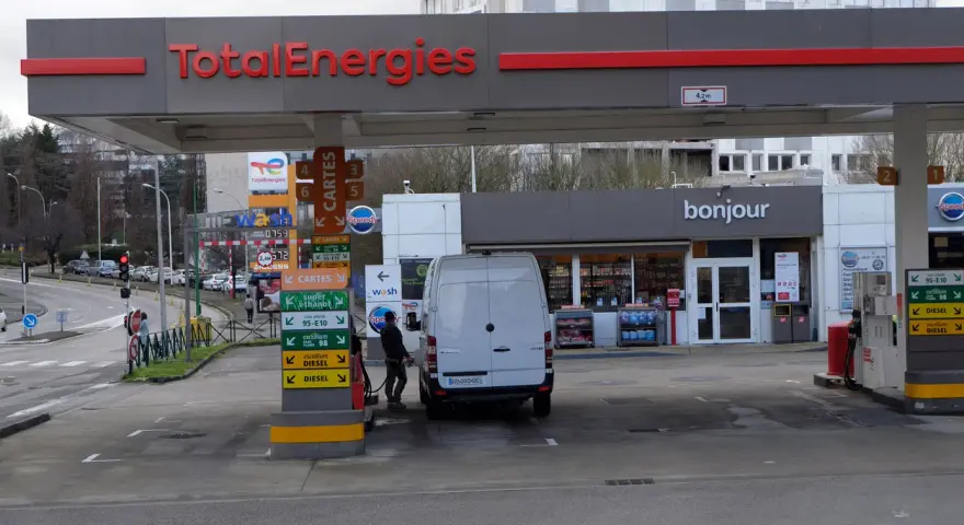 Station TotalEnergies avec une camionnette blanche en cours de ravitaillement. Panneaux de prix et de services.