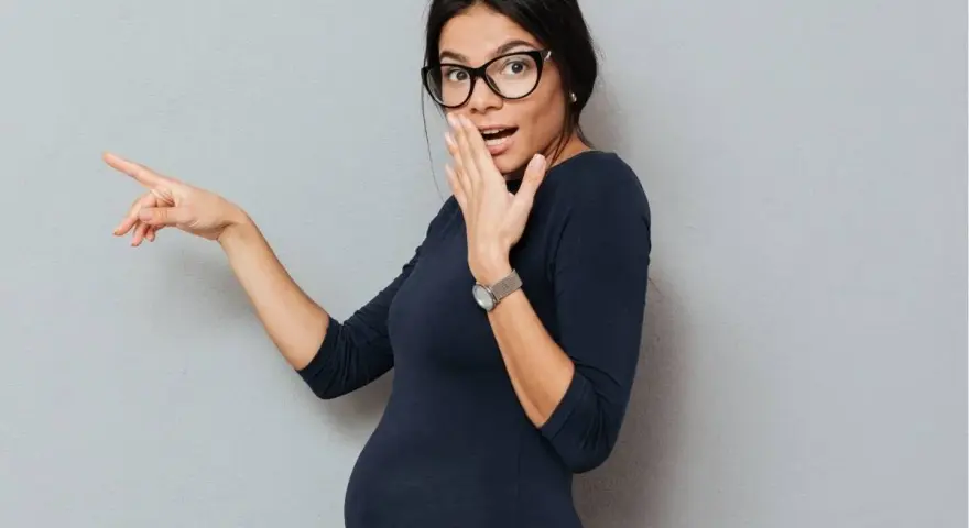 Donna in gravidanza con occhiali indica sorpresa, coprendosi la bocca con una mano.