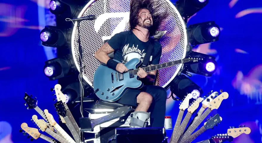 Dave Grohl z zespołem na scenie, szaleje z gitarą. Wokół niego mnóstwo gitar, a w tle wielkie logo i światła.