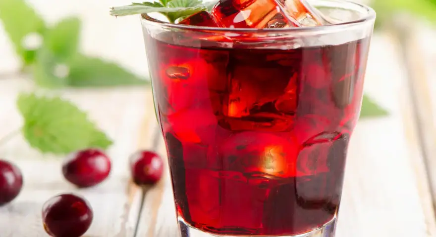 Orzeźwiający drink z żurawiną, lodem i listkiem mięty, otoczony kilkoma owocami żurawiny na drewnianym tle.
