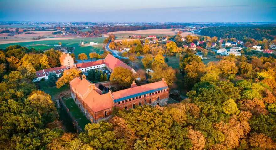 Zamek Bierzgłowski otoczony jesiennym lasem, z czerwonymi dachami i ceglanymi murami, w blasku zachodzącego słońca.