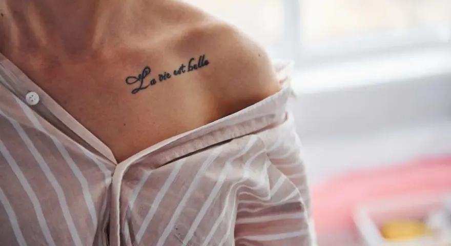 Delikatny tatuaż napis na obojczyku: "La vie est belle". Kobieta w pasiastym, rozpiętym dekoltem ubraniu.