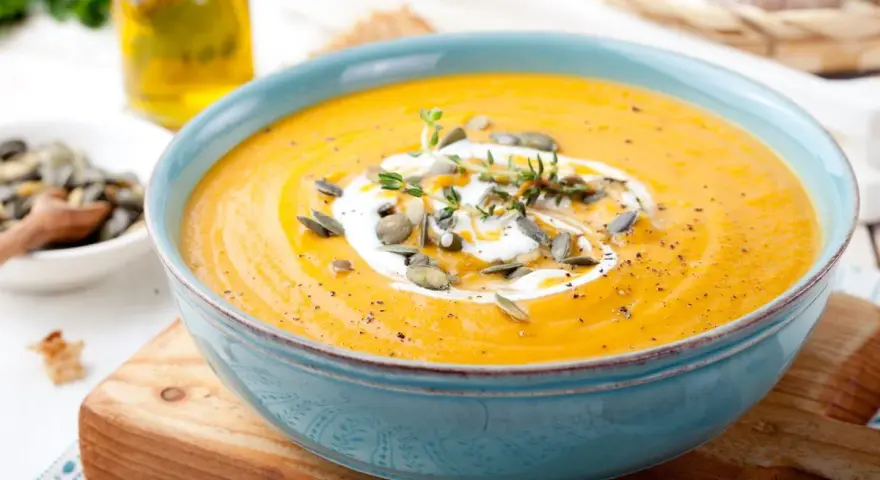 Vellutata di zucca cremosa, guarnita con panna, semi di zucca e timo, servita in una ciotola azzurra su un tagliere di legno.