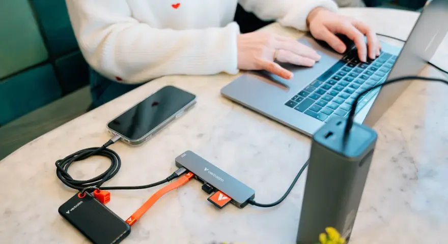 En person använder en laptop med en hubb ansluten till en telefon, extern hårddisk och powerbank. Hubben har usb 3.2-portar.