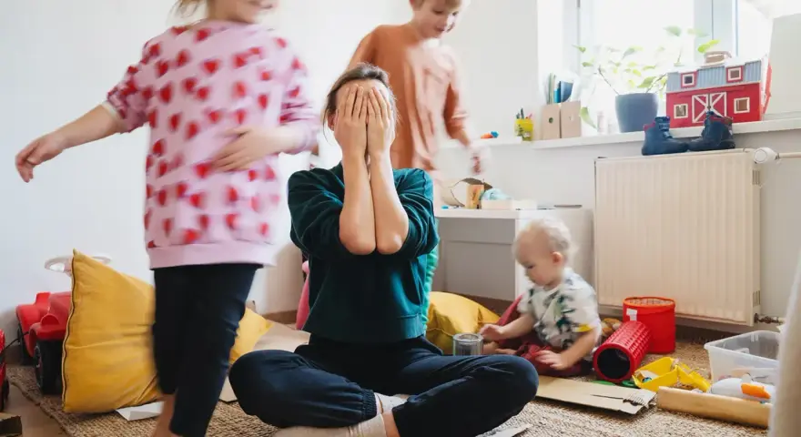 Chaos im Kinderzimmer. Eine Mutter versteckt ihr Gesicht in den Händen, während ihre Kinder spielen. Trotz des Trubels ist die liebe zu kindern spürbar.