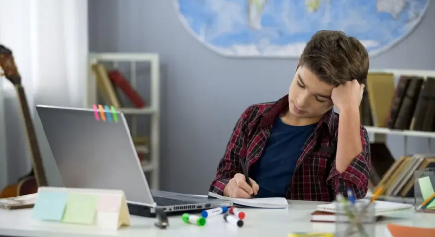 Ein Junge sitzt frustriert am Schreibtisch und macht seine hausaufgaben. Ein Laptop und bunte Stifte liegen bereit.