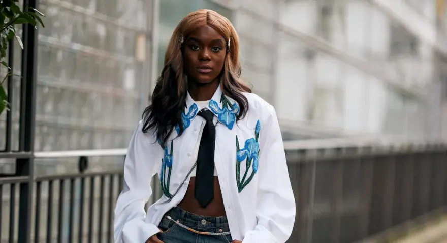 Modella con jeans baggy, un capo che simboleggia libertà e stile informale, indossa una camicia bianca con ricami floreali e cravatta.