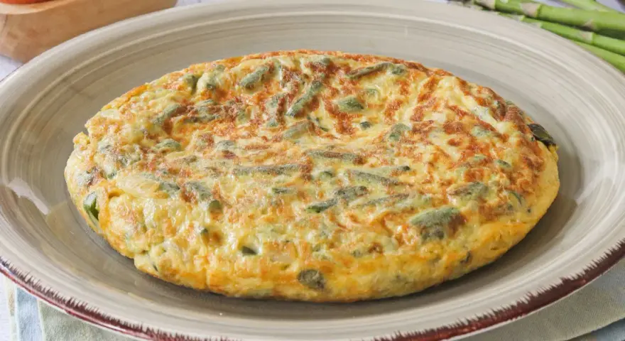 Tortilla de esparragos dorada y esponjosa, con trozos verdes de espárragos integrados en el huevo.