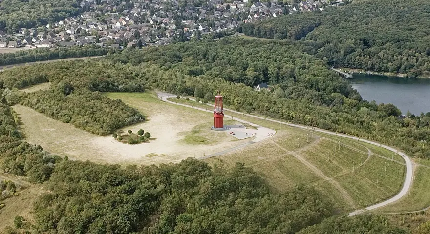 Luftaufnahme der Halde Rheinpreußen mit rotem Geleucht, umgeben von Wald und einem See.