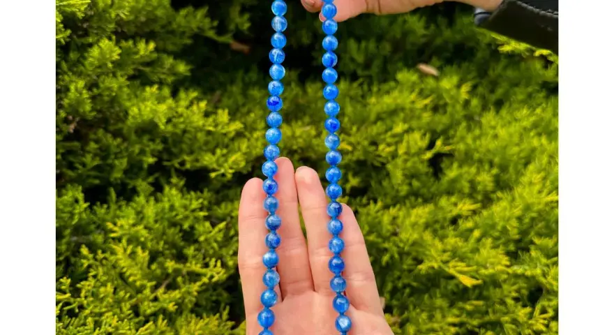 Collana di cianite blu, con perle luminose che catturano la luce, tenuta tra le mani su uno sfondo verde.