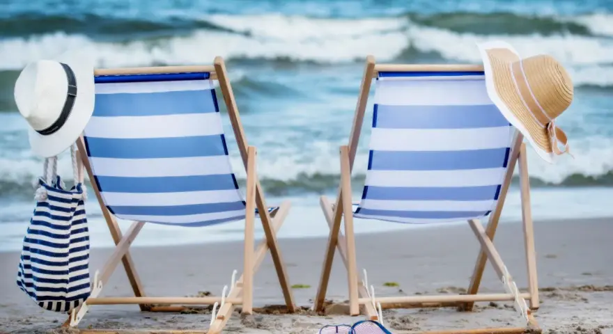 Dwa leżaki na plaży, gotowe na idealny weekend nad morzem. W tle fale, a obok kapelusze i torba plażowa.