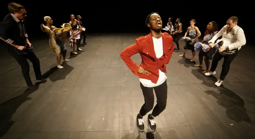 Un danseur afro 93, en veste rouge, mène une troupe sur scène. Les danseurs, aux tenues variées, exécutent des mouvements synchronisés.