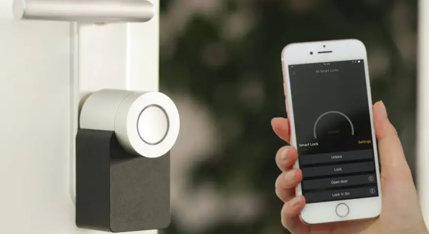 Mano sostiene un iPhone que muestra una app para controlar un cerrojo inteligente. La puerta tiene un diseño moderno.