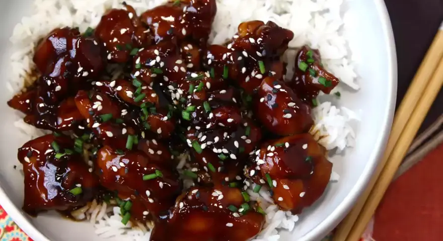Trozos de pollo teriyaki glaseados y brillantes, cubiertos con semillas de sésamo y cebollino, servidos sobre una cama de arroz blanco.