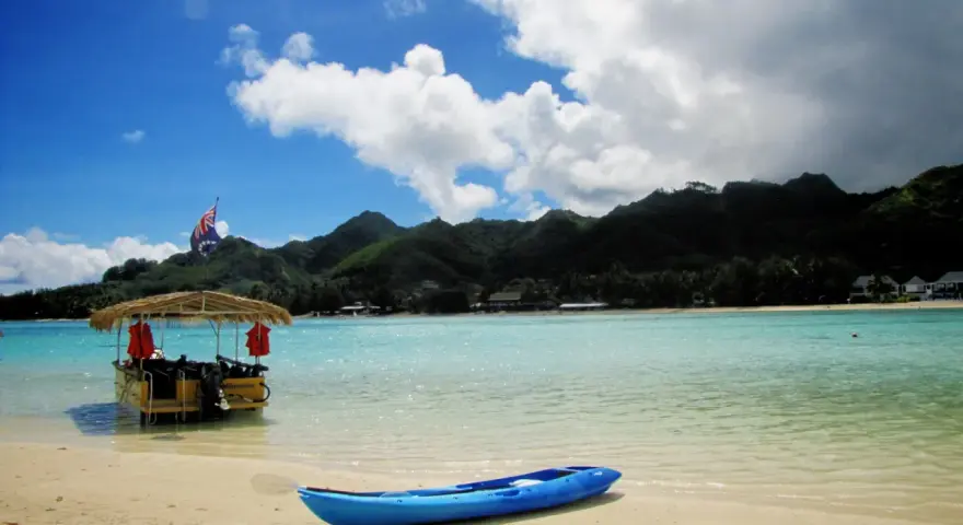 A blue kayak rests on a sandy beach in the Cook Islands, with a thatched-roof boat and lush green mountains in the background.