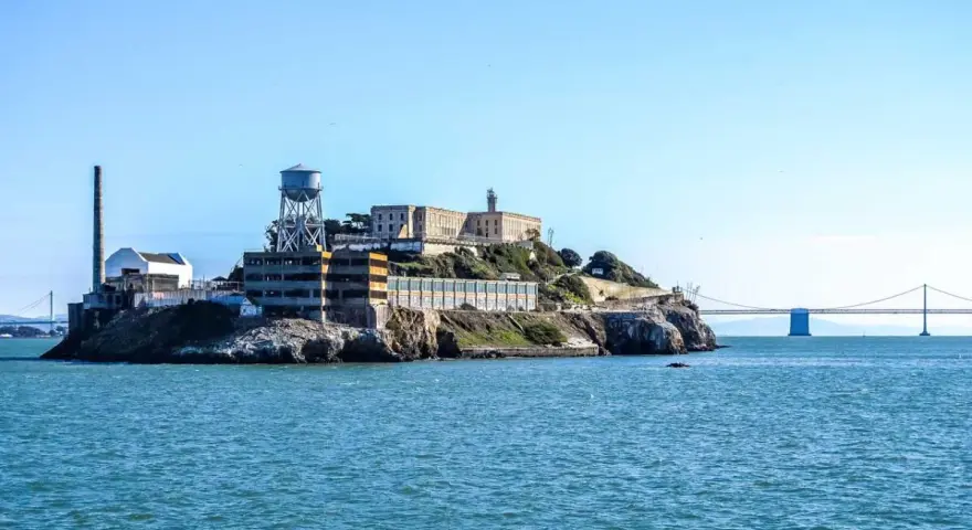 Słynne więzienie Alcatraz na skalistej wyspie, z charakterystyczną wieżą ciśnień i mostem w tle.