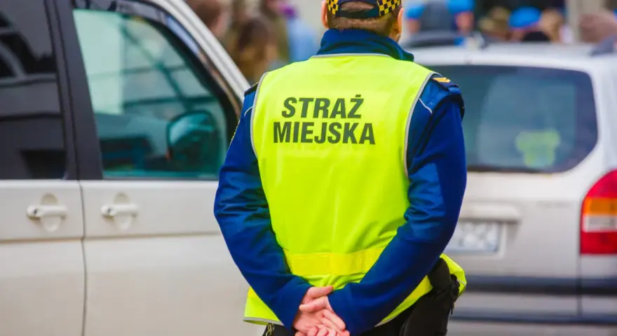Strażnik miejski w kamizelce odblaskowej stoi obok samochodu.