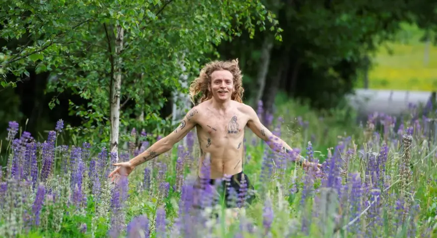 En man med lockigt hår och tatueringar står i en äng full av lila lupiner. Han ser ut att njuta av naturen, kanske som en del av ett avsnitt av "spring snyggt podden".