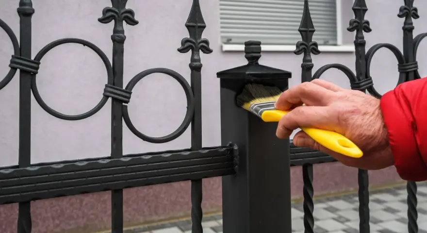 Ręka w czerwonej kurtce maluje pędzlem czarną, kutą balustradę balkonu w bloku.