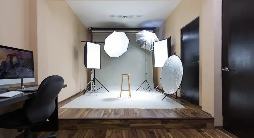Domowe studio fotograficzne z oświetleniem, statywami, blendą i krzesłem. Po lewej komputer z monitorem.
