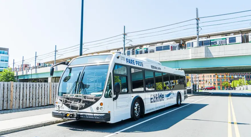 A white EZRide bus, offering fare-free transit from Boston to Cambridge, MA, drives past an elevated train station.