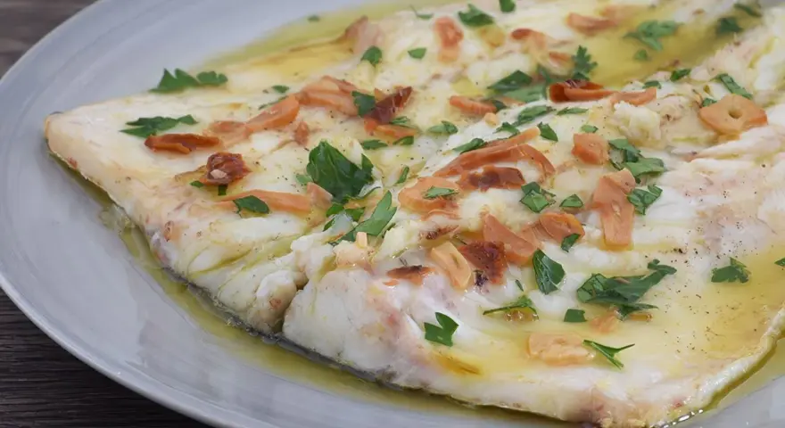 Pescado blanco cocinado en aceite con ajo laminado y perejil picado, servido en plato gris.