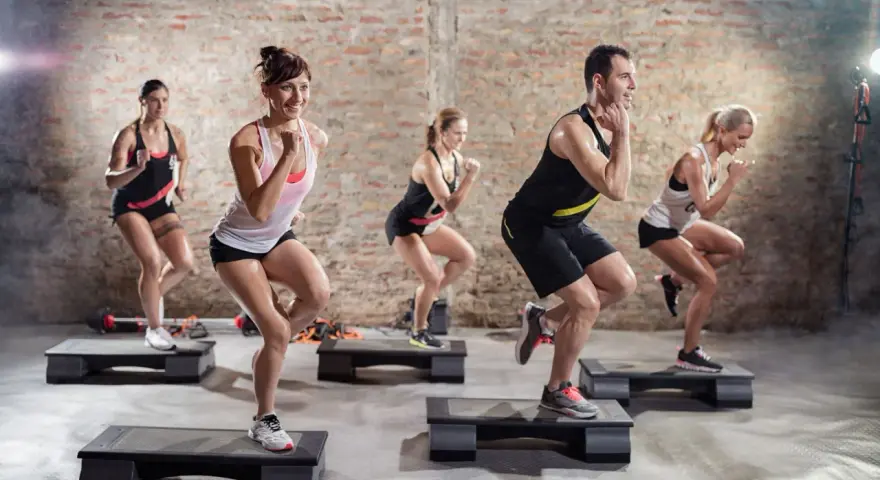 Un groupe de personnes participe à un cours step, effectuant des mouvements dynamiques sur des plateformes d'exercice dans une salle de sport.