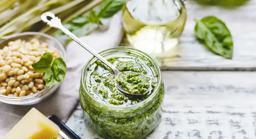 Un barattolo pieno di profumato pesto alla genovese, con pinoli, basilico e olio d'oliva pronti per essere mescolati.