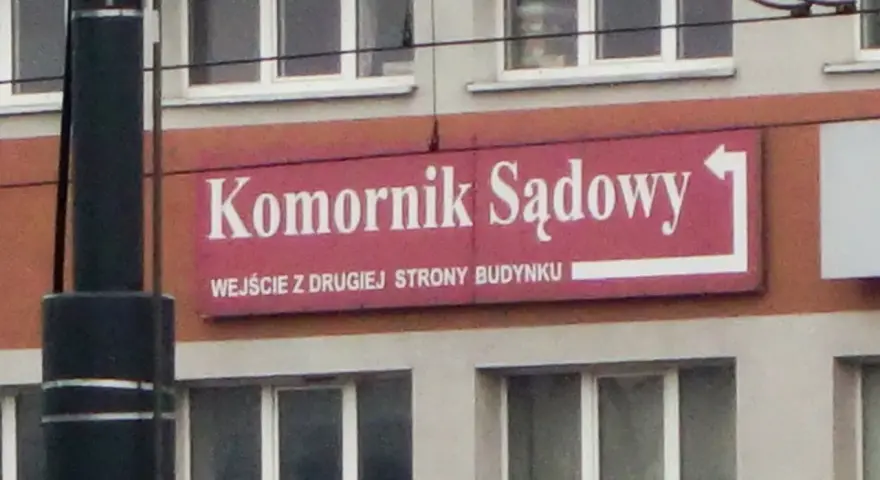 Tablica "Komornik Sądowy" na budynku. Wskazuje wejście z drugiej strony.