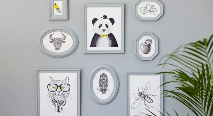 En samling gråa ramar med djur och föremål. En panda med fluga, en tjur, en katt med glasögon, en uggla, en bi, ett äpple och en cykel. Någon har valt att måla ramar för att skapa en personlig vägg.