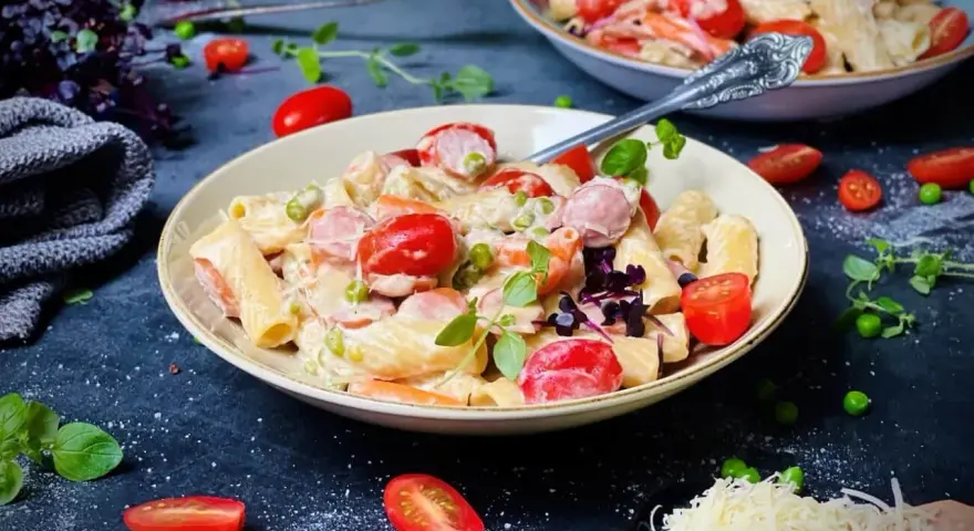 Cremige One Pot Pasta mit Würstchen, Erbsen und Kirschtomaten, garniert mit frischen Kräutern und geriebenem Käse.