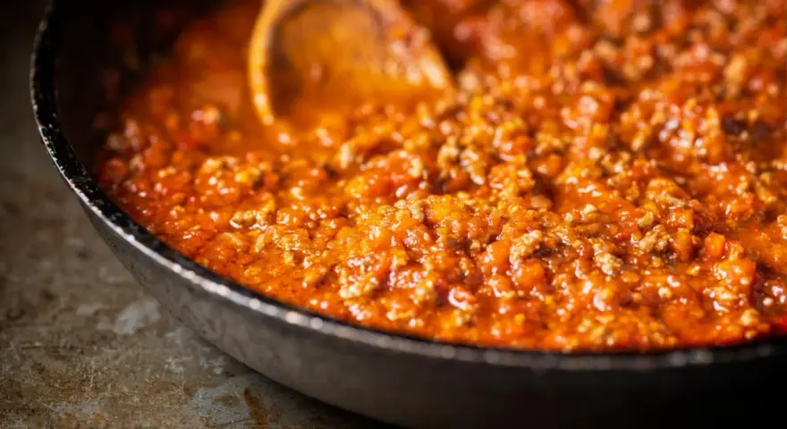 Ein Holzlöffel rührt in einer Pfanne mit köchelndem Sos bolognese.