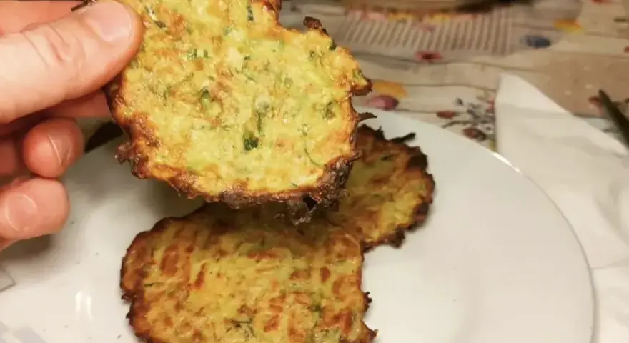 Una mano tiene una frittella di zucchine dorata e croccante. Sul piatto, altre frittelle di zucchine invitano ad essere gustate.