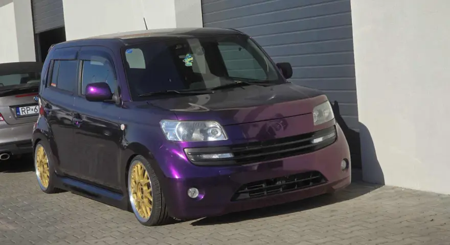 Fioletowy Daihatsu Materia z błyszczącymi złotymi felgami, zaparkowany przed garażem.