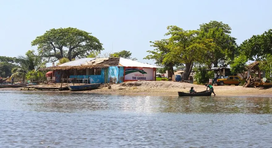 Nad rzeką w Gambii, przy brzegu z łodziami i budynkami, widać tablicę "Welcome to THE GAMBIA".