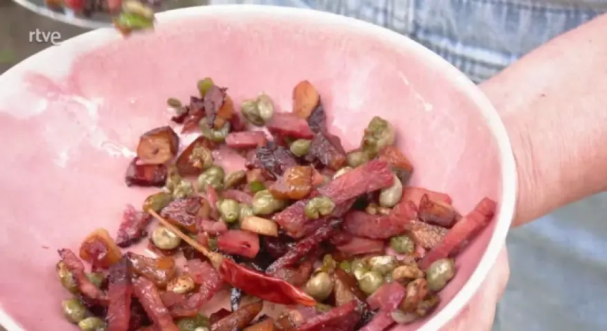 Un plato rosa lleno de habas con jamón, guisantes y un chile rojo seco.
