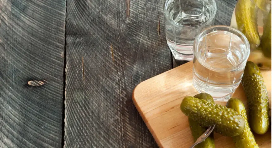 Zwei Gläser Wodka und eingelegte Gurken auf einem Holzbrett. Perfekt für alle, die Wodka selber machen wollen.