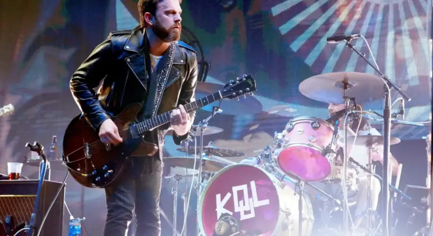 Gitarzysta Kings of Leon Polska w skórzanej kurtce gra na scenie. W tle perkusja z logo KQL.
