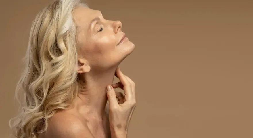 Kobieta z blond włosami i zamkniętymi oczami delikatnie dotyka szyi. Jej skóra w okresie menopauzy jest widoczna, podkreślając naturalne piękno.