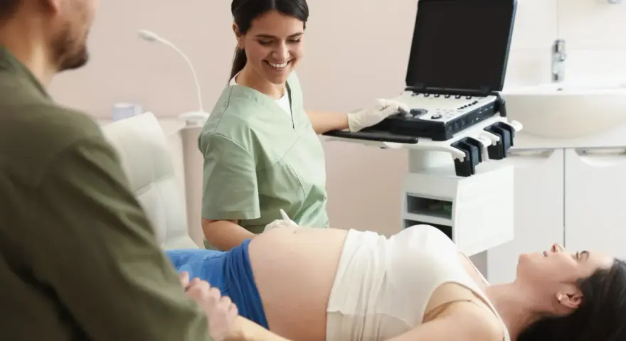Uśmiechnięta lekarka wykonuje badanie wolnego płodowego dna u ciężarnej pacjentki, podczas gdy partner wspiera ją, trzymając za rękę.