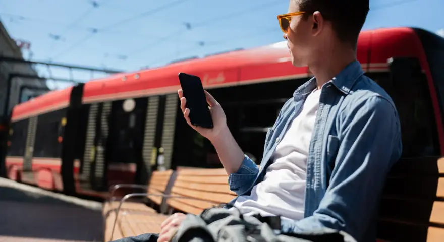 Mężczyzna w okularach siedzi na ławce i trzyma telefon. W tle czerwony tramwaj.