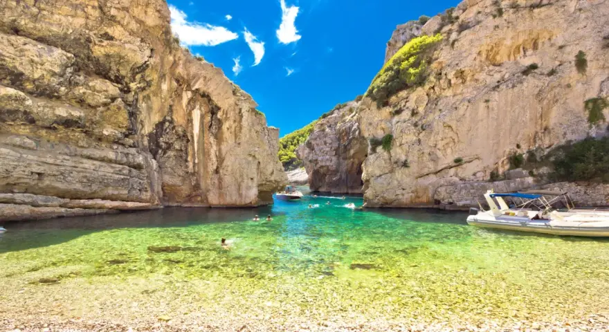 Spiaggia di ciottoli e acque cristalline tra le rocce dell'isola di Lissa, con persone che nuotano e barche.