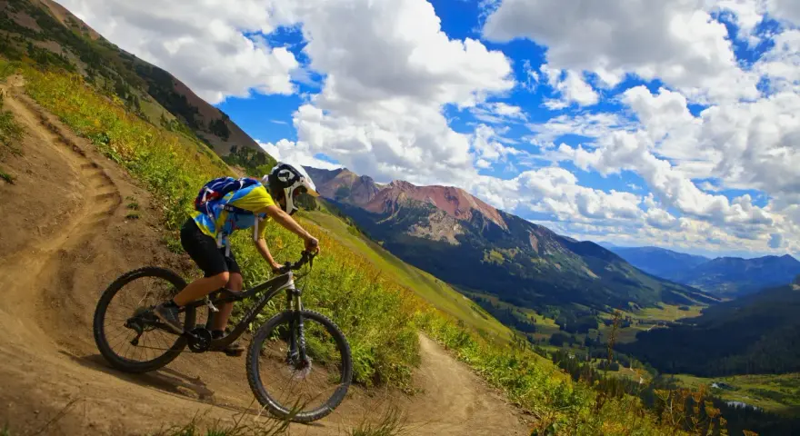 A mountain biker navigates a winding trail, enjoying the breathtaking views of cross country mountain bike trails winding through a lush valley and rugged peaks under a dramatic sky.
