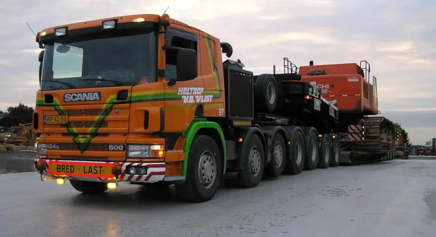 Pomarańczowy Scania z naczepą przewozi koparkę. Transport ponadgabarytowy wymaga specjalnych godzin jazdy.
